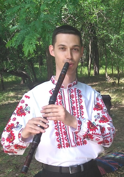 Photo of Kiril Belezhkov in an embroidered shirt, playing the kaval in a forest clearing.
