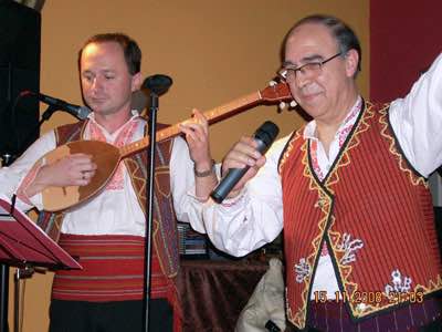 Photo of Stefče Stojkovski in costume playing the tambura on stage, next to Dragi Spasovski dancing in costume